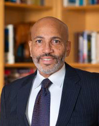 A person in a suit and tie stands in front of a bookshelf.